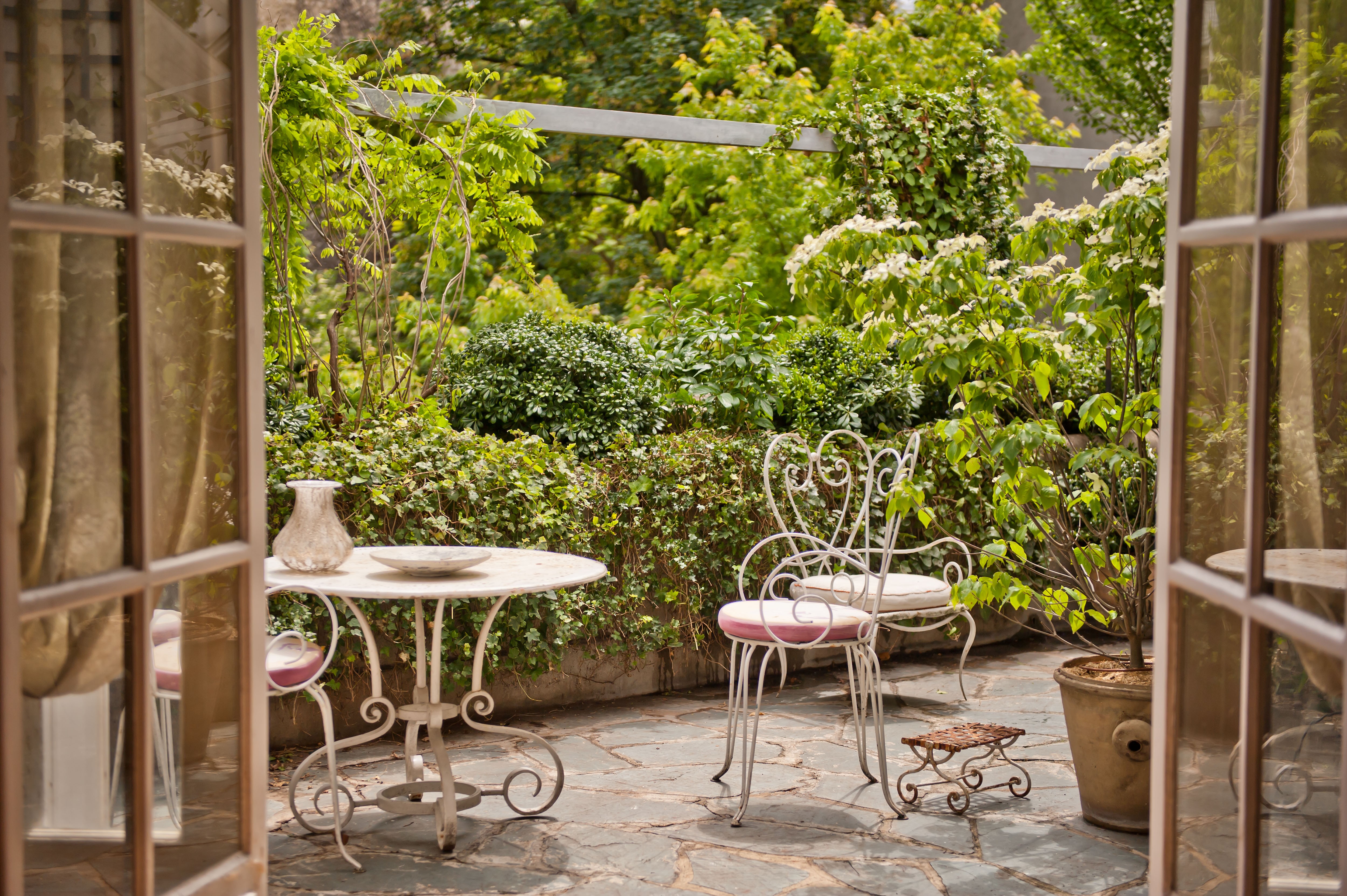Depuis une grande fenêtre ouverte vue sur une terrasse en ardoise d'un hôtel particulier parisien avec meubles de jardin en fer forgé blanc