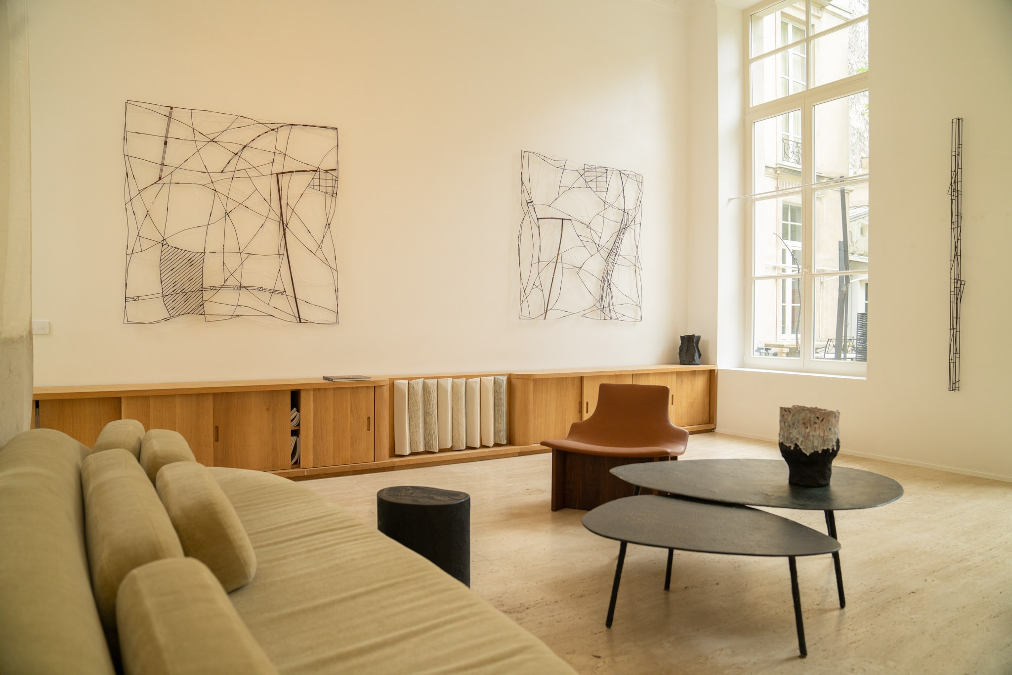 Intérieur lumineux et épuré avec un canapé blanc, un sol beige, une table basse aux formes organiques et une œuvre d&#039;art filaire noire accrochée au mur.