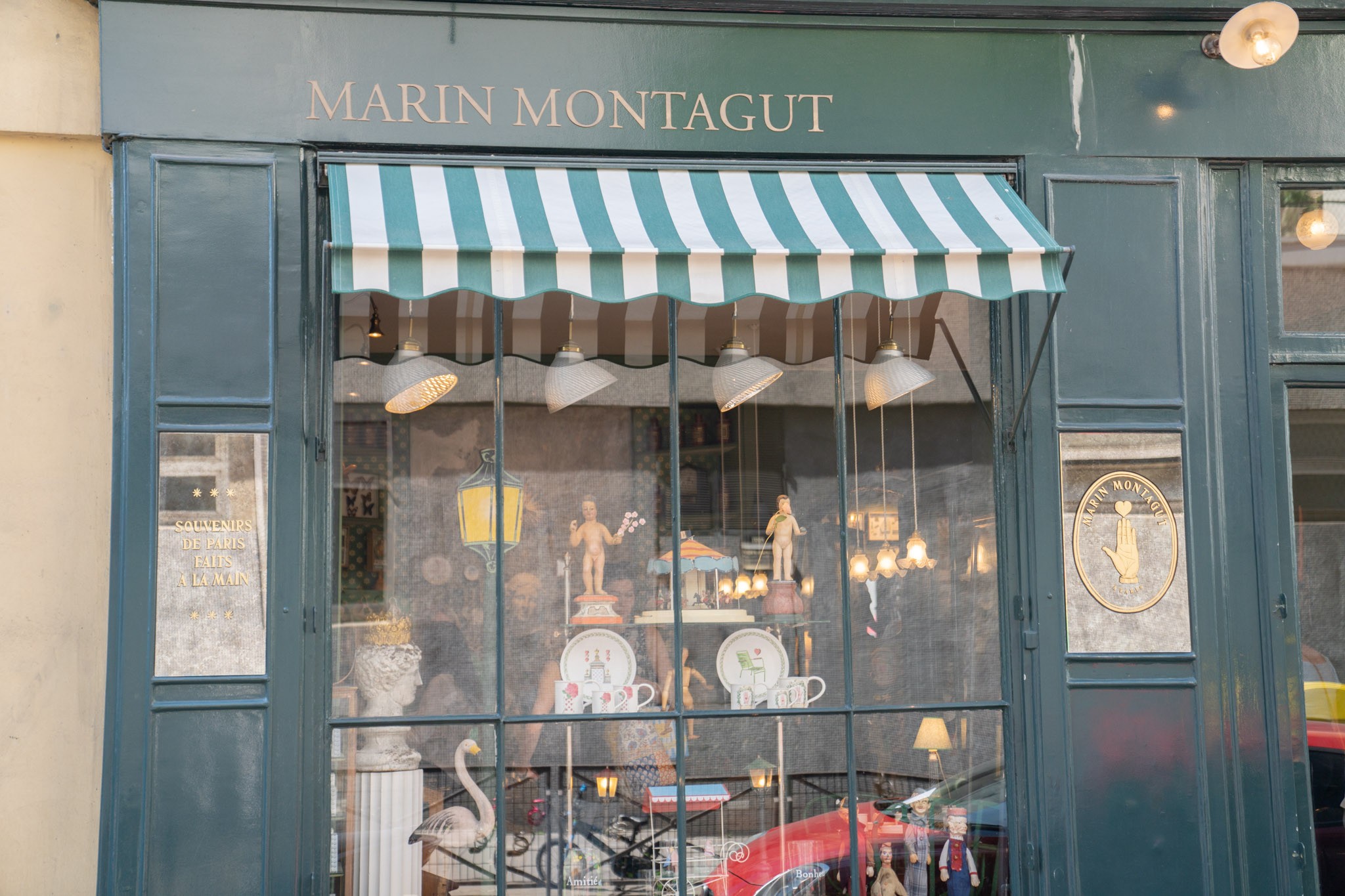 Devanture de la boutique de Marin Montagut à Paris, avec un store à rayures vertes et blanches. La vitrine expose des objets d'antiquaire.