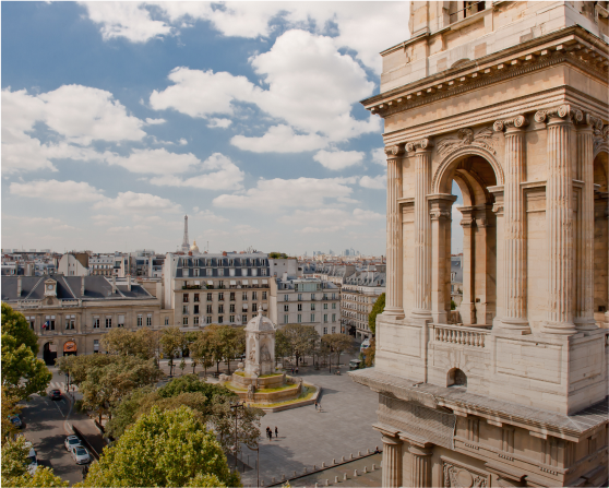 Vue aérienne de la place Saint-Sulpice, capturant le parvis de l'église, le clocher, la fontaine, les immeubles haussmanniens et les grands arbres. La Tour Eiffel et les Invalides apparaissent en arrière-plan.
