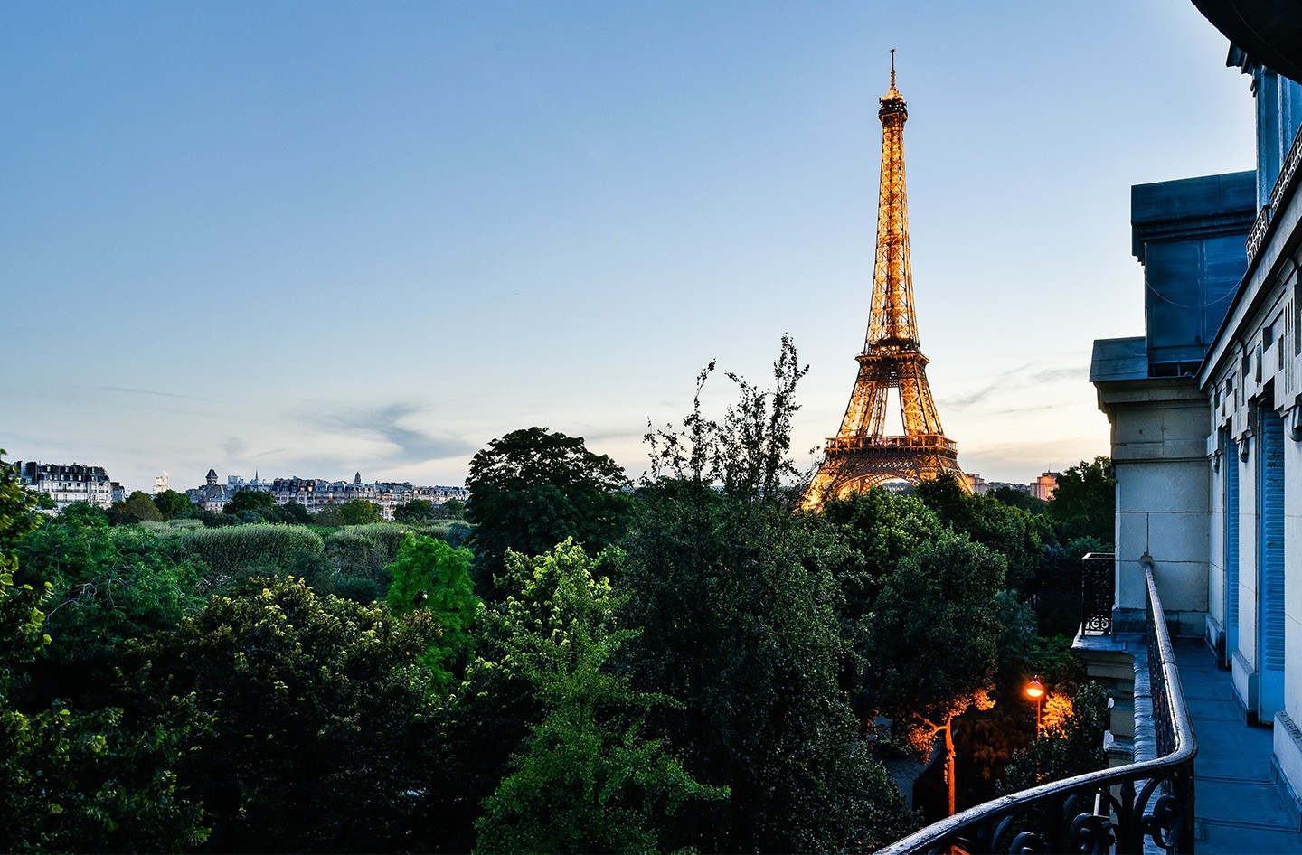Vue depuis un balcon parisien en fin de soirée, montrant un bâtiment haussmannien avec son toit en zinc. La lumière douce de la fin de journée éclaire la cime des arbres et la Tour Eiffel illuminée.